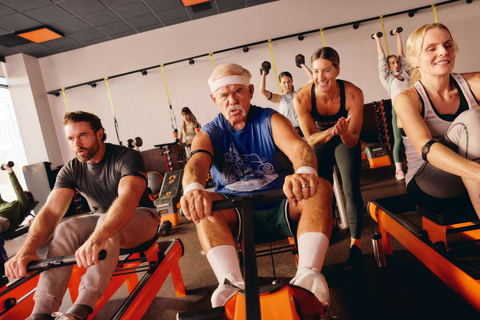 A diverse group of people work out on rowing machines in a brightly lit fitness studio, with a coach encouraging them from behind.