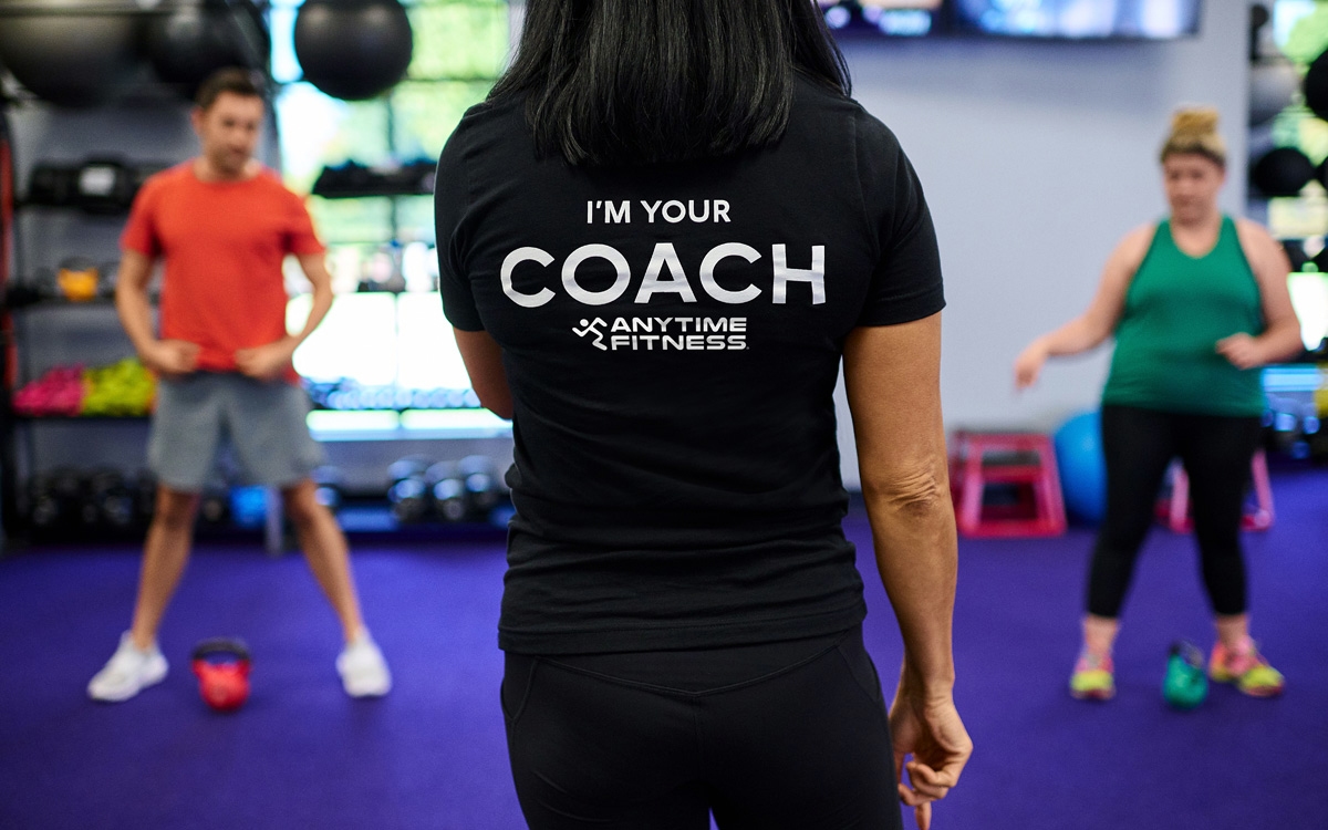A fitness coach wearing a shirt that reads “I’m Your Coach – Anytime Fitness” stands in front of two participants during a workout session in a gym.