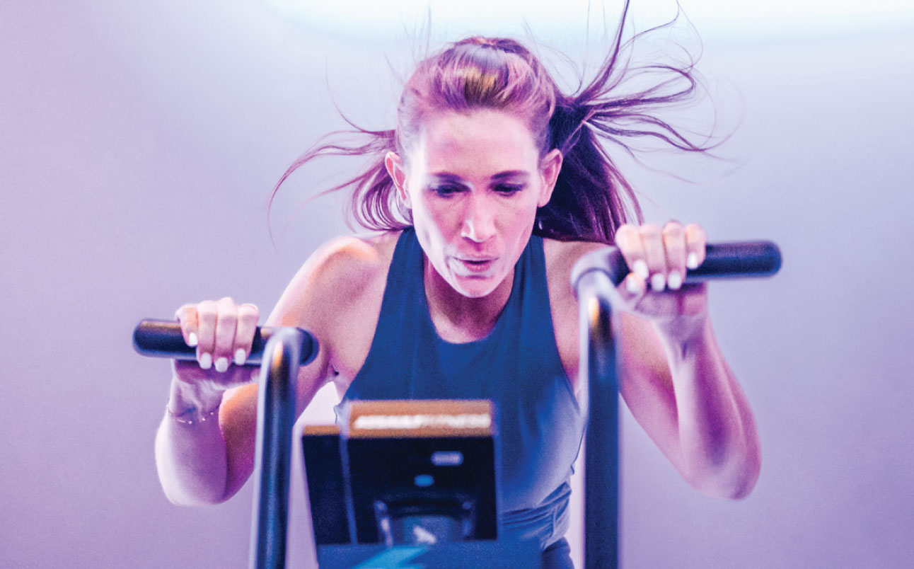 A woman intensely focused during a high-energy workout on an air bike, her hair flying back and muscles engaged under dramatic lighting in a fitness studio.