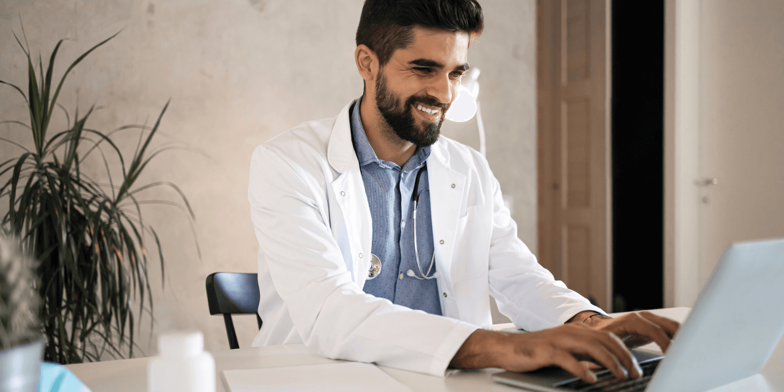 Un médico varón de piel de tono medio y cabello oscuro está sentado con una camisa azul y una bata de laboratorio con un estetoscopio alrededor del cuello, escribiendo en una computadora y sonriendo