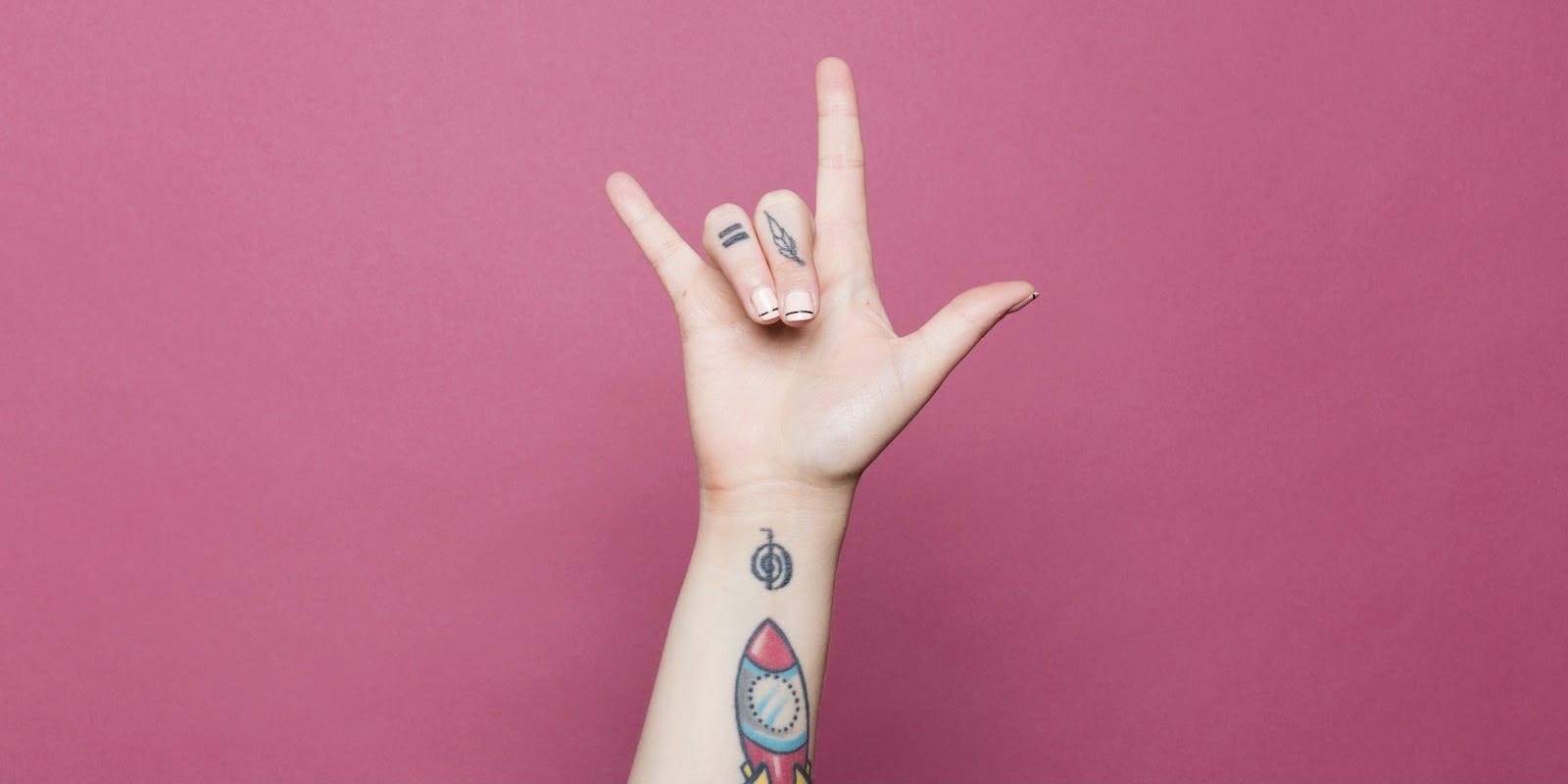 Un antebrazo tatuado se extiende sobre un fondo rosa brillante con una mano que deletrea "Te amo" en lengua de signos.