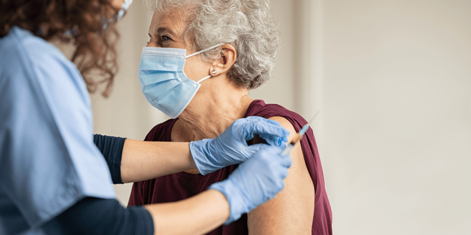 Un técnico médico, de pelo rizado y vestido con camisa oscura de manga larga y bata, le administra una inyección en la parte superior del brazo a una mujer mayor de edad, de pelo corto y gris, que lleva puesta una máscara médica y una camiseta roja.