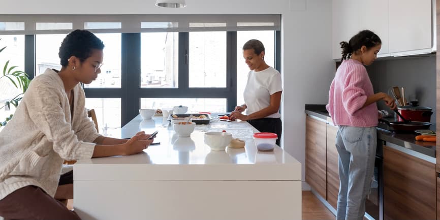 A photograph of a large kitchen with a Black mother and teenage children, one who is cooking at the stove in jeans and a pink sweatshirt and the other who is sitting at the counter wearing a white button down shirt and looking into their phone.