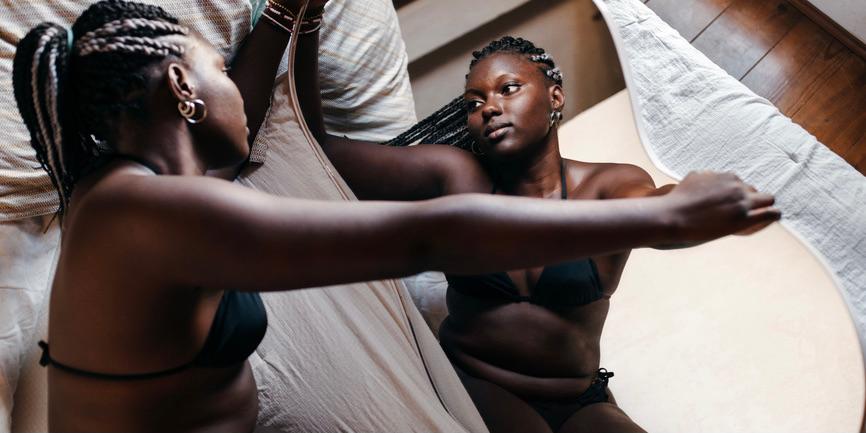 A color photograph of a young Black woman with a medium sized body in bed wearing black underwear and a bra and looks into a mirror, holding a sheet loosely over her head.