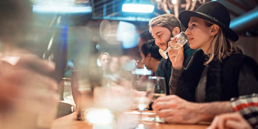 A color photograph of a bar, where a young white woman with blond hair wearing a black hat and sweater takes a sip of wine next to a young white man doing the same.