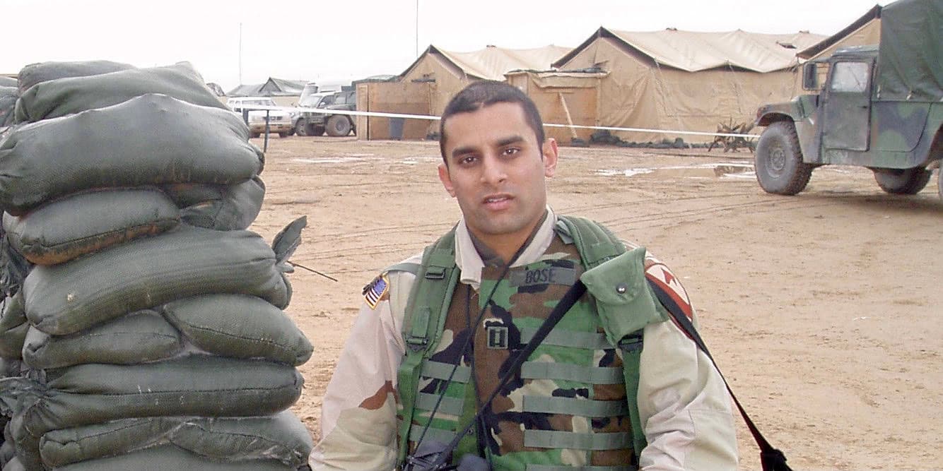 Una fotografía del Dr. Sudip Bose con su uniforme militar, de pie frente a una fila de tiendas de campaña y un camión militar.