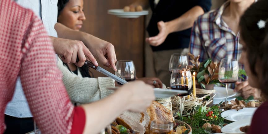 A close-up color photograph of a dining room table full of food with various people passing dishes to each other around it.