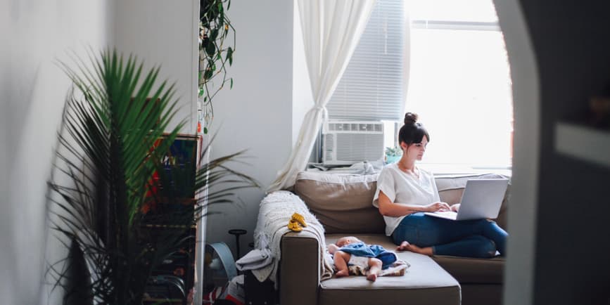 Una fotografía en color de una mujer blanca con el pelo largo recogido, trabajando en una computadora mientras está sentada en su sala de estar con un bebé descansando en el sofá de al lado.