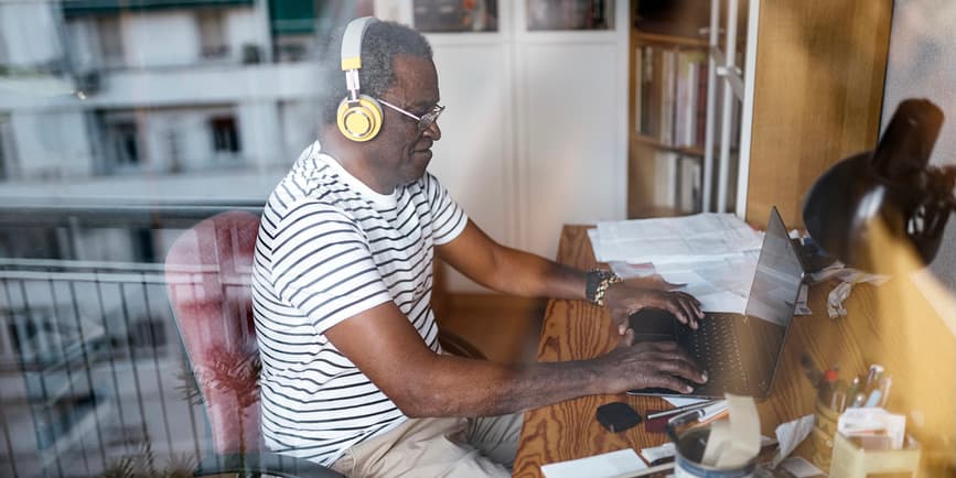 A color photograph taken through a window shows an older Black man with short white hair wearing a striped t-shirt and headphones sitting and typing at a computer.