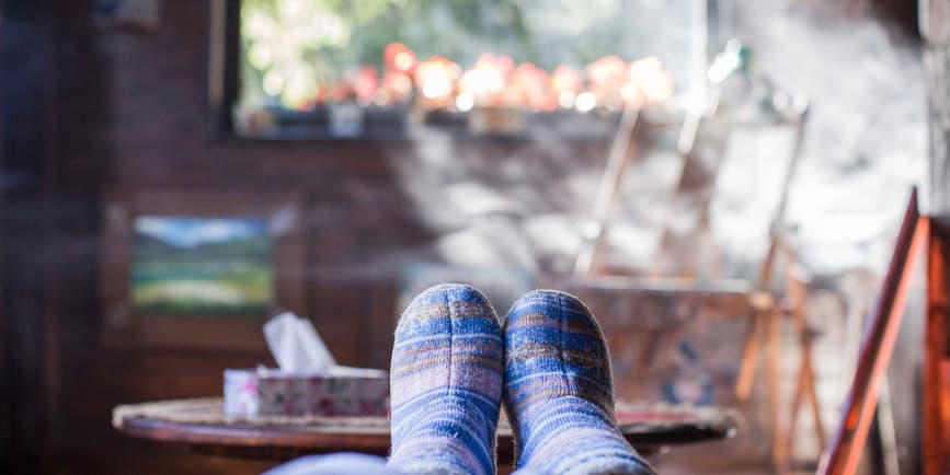 Una fotografía interior en color de una alegre escena invernal, con pies resbaladizos posados sobre una mesa de café frente a una ventana brillante y brumosa con luces navideñas en el exterior.