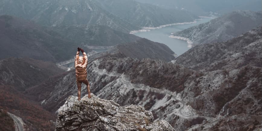 Un colorido paisaje exterior de una cadena montañosa con un río que la atraviesa. En primer plano, una mujer está de espaldas a la cámara, con pantalones marrones y una chaqueta dorada brillante, levantando los brazos hacia el cielo.