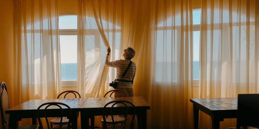 Una fotografía en color de interiores de un turista masculino mayor y blanco con una cámara alrededor del cuello, de pie junto a una ventana entre mesas de comedor vacías, abriendo cortinas amarillas de gasa para ver el agua y el cielo.