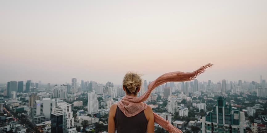 Una fotografía en color al aire libre de una mujer blanca con el pelo rubio recogido en un moño y un pañuelo rosa que fluye con el viento. Está de pie de espaldas a la cámara, con una amplia escena de la ciudad de Nueva York y un cielo rosa por delante.