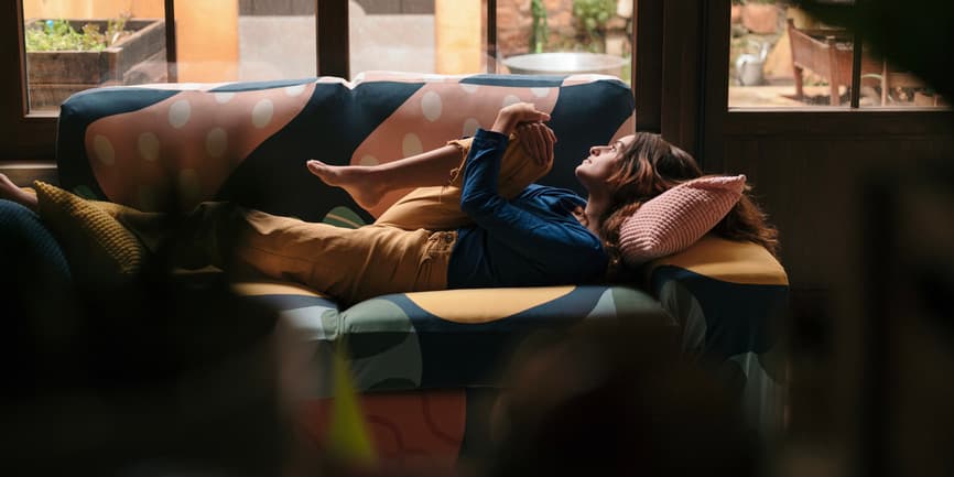 An indoor photograph of a white woman with long blond hair wearing lounge pants and a long-sleeve shirt, laying on a couch with one knee pulled up, looking out a large window