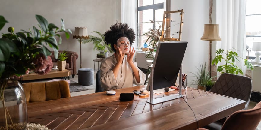 A color indoor photograph of a young Black woman wearing a tan robe with afro hair pulled back with a headband. She's sitting at a table in a sunlit living room surrounded by plants, looking into a mirror while putting on a sheet facial mask.