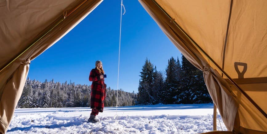 A color outdoor photograph taken from the inside of a tent looking out, where a young white woman wearing a red plaid robe stands in the snow against a blue sky and fir trees, drinking a cup of coffee.