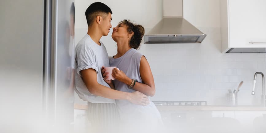 Una fotografía interior en color de una pareja en una cocina blanca y de acero. Un joven asiático de pelo corto con una camiseta gris abraza a una joven de piel morena, que también lleva un té gris de manga corta, que sostiene una taza de café.