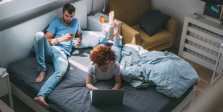 A color photograph looking down on a young white couple laying on top of their bed. They're both wearing jeans and a t-shirt. She's typing on a computer and he's looking down into his phone.