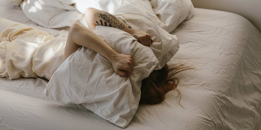 Una mujer blanca está acostada boca arriba en la cama con una almohada sobre la cabeza, lo que significa malestar
