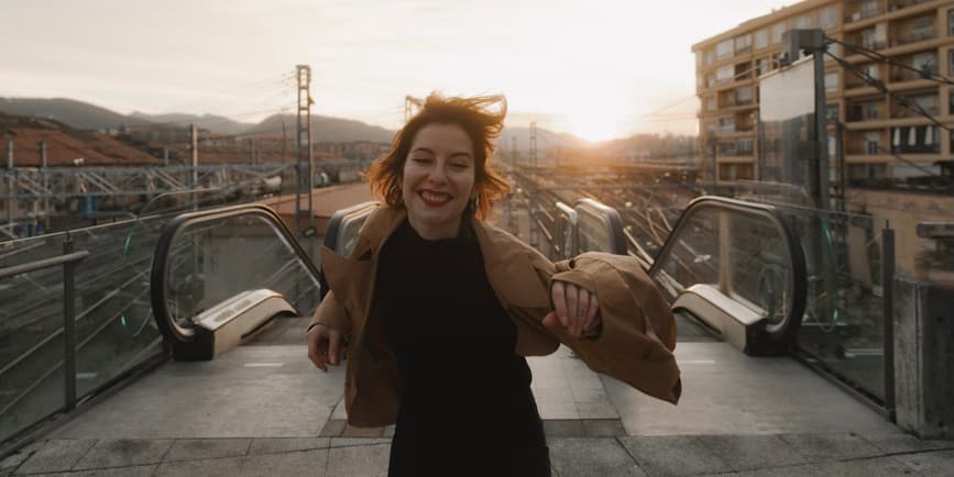 An outdoor color photograph of a white woman with short red hair, wearing red lipstick, a brown coat and a black shirt. She's standing outside in front of a city scene on a train platform, smiling big as she moves towards the camera.