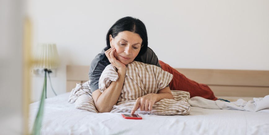 Una mujer de mediana edad con cabello oscuro está acostada en la cama mirando un teléfono en una funda roja
