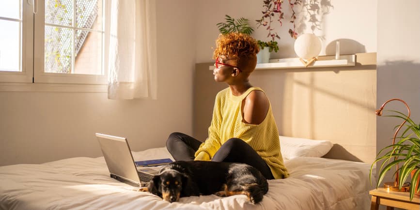 Una fotografía interior en color de una joven negra con cabello castaño con un suéter amarillo y pantalones negros. Se sientan con las piernas cruzadas en una cama frente a un portátil con un perro durmiendo a su lado, mirando por la soleada ventana.