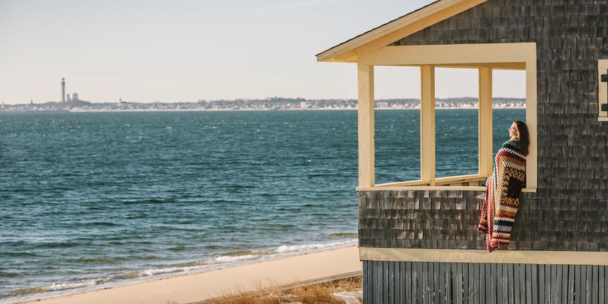 A color outdoor photograph of a grey-shingled house next to a large beach and blue water, where a white woman sits on the porch looking out at the ocean, a colorful quilt wrapped around her.