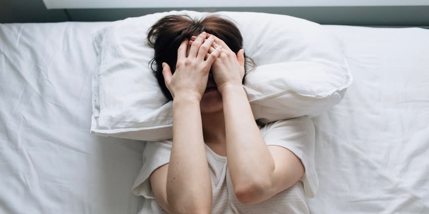 una mujer blanca con una camiseta blanca se acuesta en la cama sobre sábanas blancas con las manos sobre la cara, incómoda