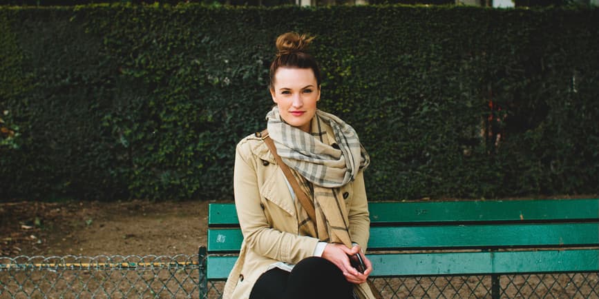 A young white woman her hair up in a knot wearing a trench coat and long scarf sits on a bench against a wall of green shrubbery in Paris France.