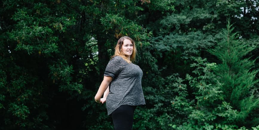 A plus-size, beautiful white woman stands stretching in workout clothes in front of a wall of green trees.