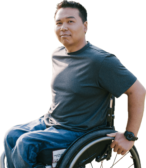 Hombre asiático en silla de ruedas, sentado al sol sobre la hierba, con las manos sobre las ruedas, sonriendo sutilmente a la cámara.