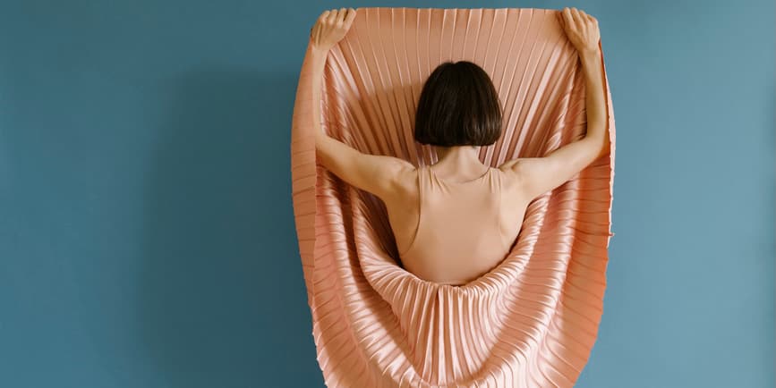 A white woman with short brown hair stands with her body turned to a bright blue wall, lifting the skirt of her pinkish pleated dress up above her head.