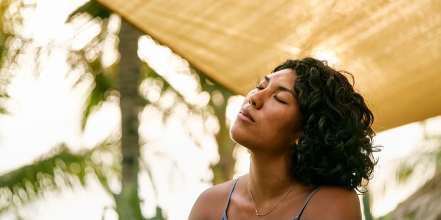 Una fotografía al aire libre de una hermosa mujer de piel morena con cabello negro y rizado. Está sentada bajo una sombrilla con palmeras al fondo, con los ojos cerrados y el rostro inclinado hacia el cielo.