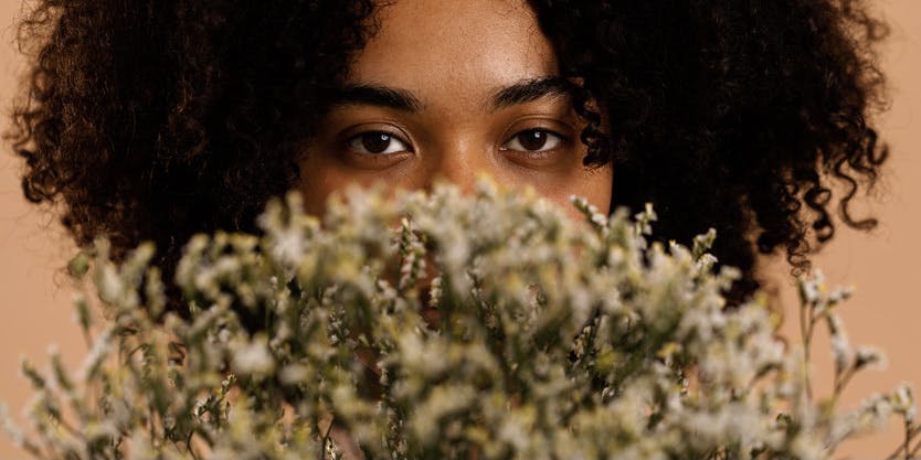 Una fotografía de primer plano de una mujer negra con cabello castaño rizado parada frente a una pared de color melocotón. Sostiene un ramo de flores silvestres cerca de su rostro, por lo que solo se ven sus ojos hinchados detrás de él.