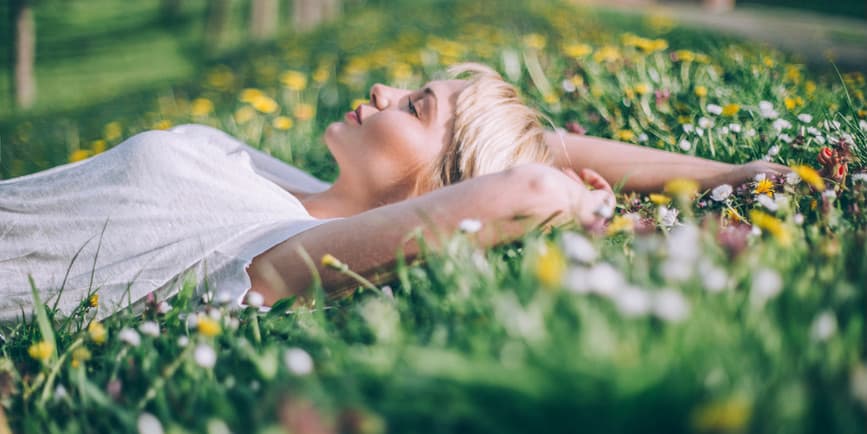 Fotografía en color de una joven blanca de pelo corto y rubio con una camiseta blanca, tumbada afuera en un campo de flores silvestres con la cara levantada hacia el cielo.