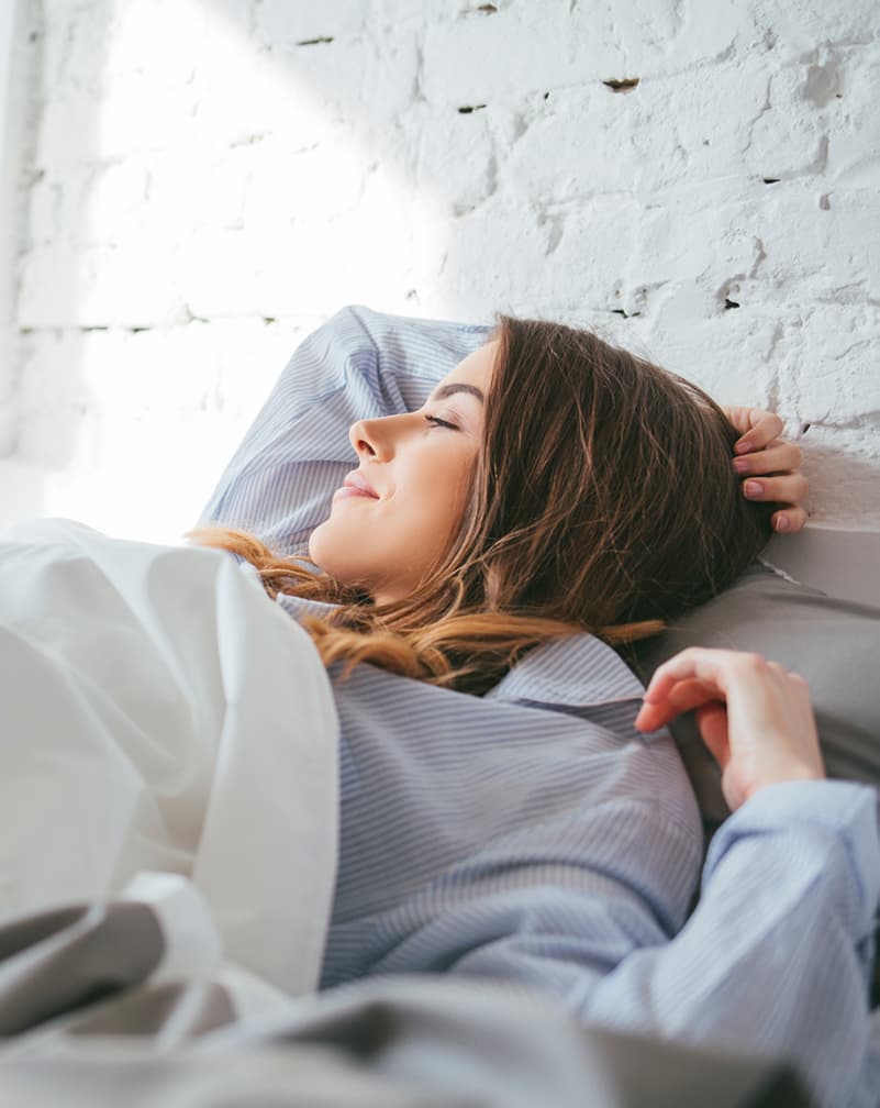 Mujer joven acostada en la cama, sonriendo hacia su ventana llena de sol con una mano sobre la cabeza