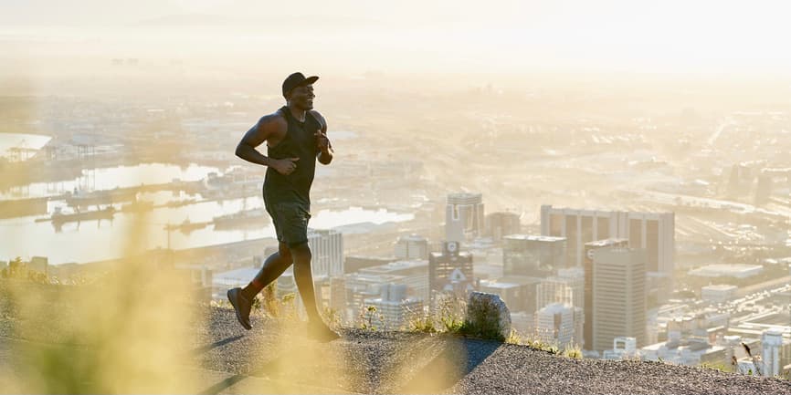 Un hombre negro en forma corre por la calle al amanecer. Está bien construido y viste ropa deportiva completamente negra mientras corre por una carretera con vistas a una ciudad al amanecer.