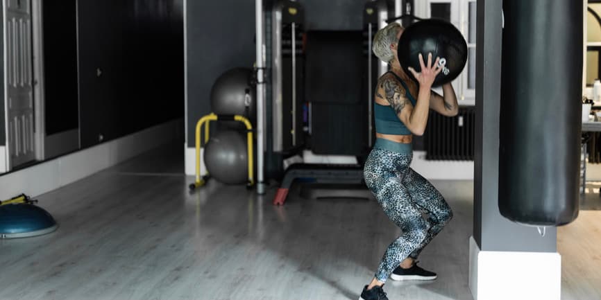 An older white woman in blue leggings and a cropped top squats with weighted balls at the gym.