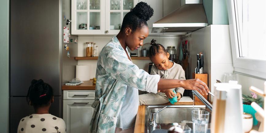 A Black woman wearing a white and blue robe and her little daughters wash dishes in a sunlit kitchen.