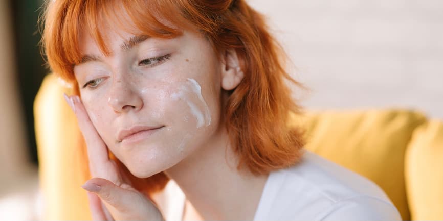 Una joven blanca de piel clara y pelo rojo corto que lleva una camiseta blanca se pone crema hidratante en la cara mientras mira fuera de cámara.