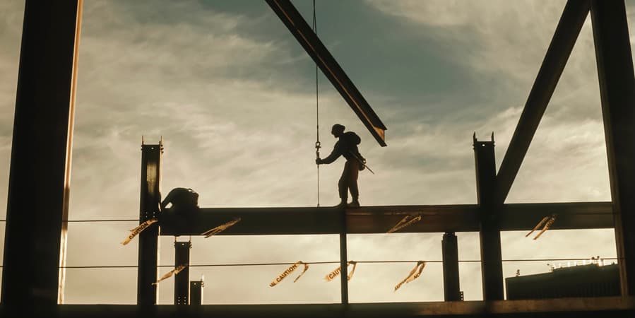 A construction worker walks on a steel rail amongst other steel scaffolding and a cloudy sky behind him.