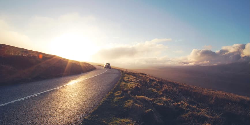 A wide outdoor photograph of a paved road surrounded by mountains and a cloudy sky, with a car driving off into the distance.