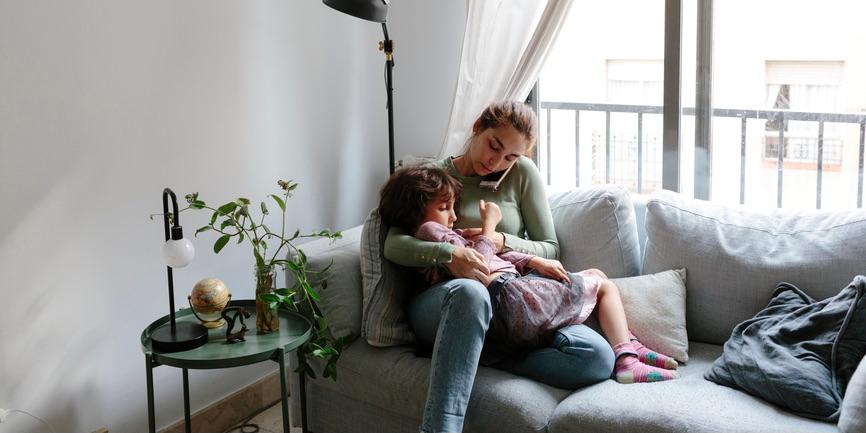 Joven morena sentada en el sofá de la sala de estar de su casa, con un niño de 6 años en su regazo. Está al teléfono haciendo una consulta con un médico. La mujer está comprobando la temperatura del niño con un termómetro.