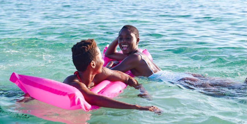 Dos jóvenes amigas africanas que ríen se aferran a una carroza mientras nadan juntas en el océano en un día soleado.