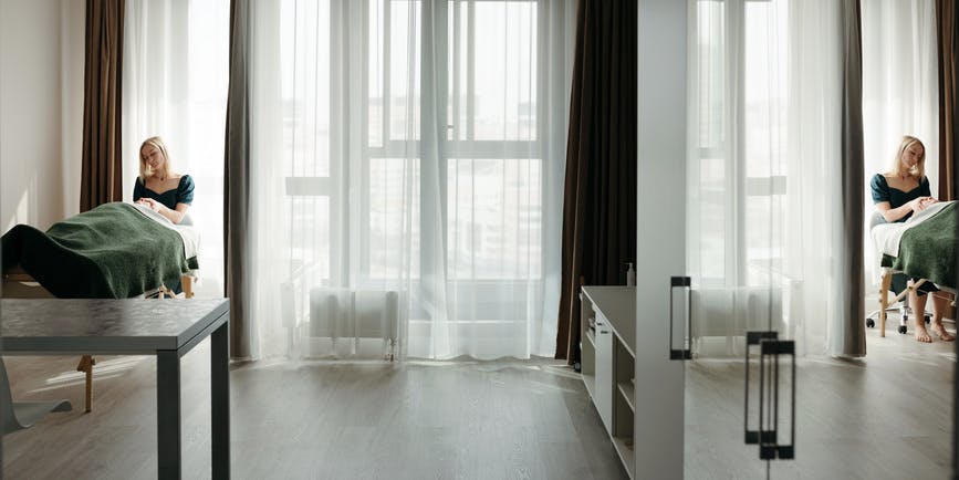 A bright, airy room where a white female massage therapists works on a client lying on a massage table under a green blanket. 