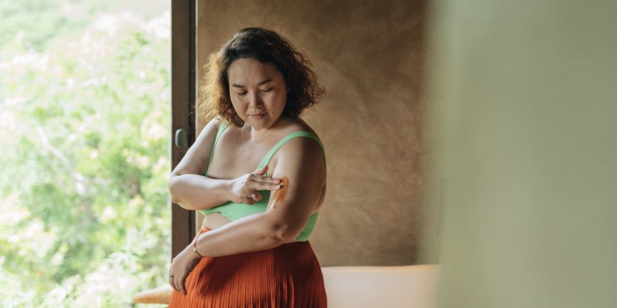 A curvy woman with brown skin and brown hair wearing a green tank top and orange skirt applies a cream to her upper arm.