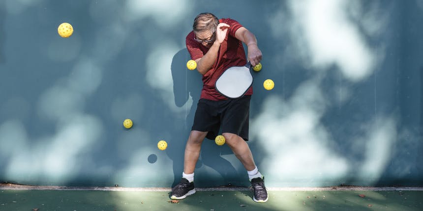 Un hombre blanco vestido con una camisa roja y pantalones cortos negros está de pie contra una pared azul con una raqueta de pickleball mientras bolas amarillas vuelan hacia él.