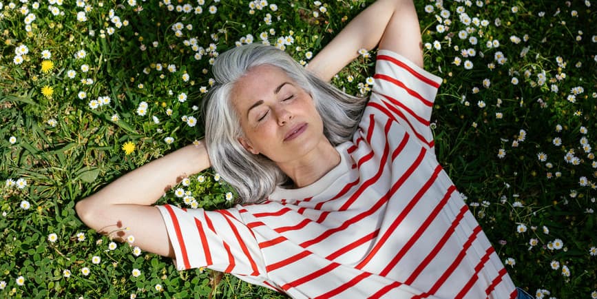 Top view of peaceful middle aged female with gray hair holding hands behind head and closing eyes while resting on blooming grass