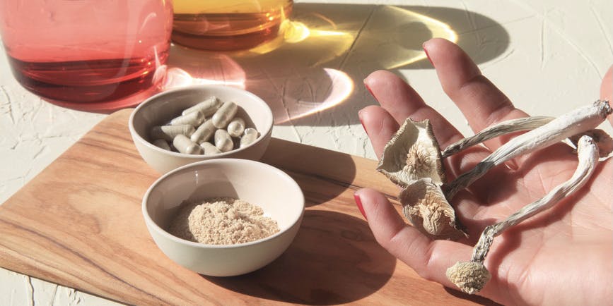 A white hand with red-painted fingernails holds a few dried psilocybin mushrooms on top of a wood cutting board. Bowls of dried mushroom powder and mushroom capsules sit nearby.