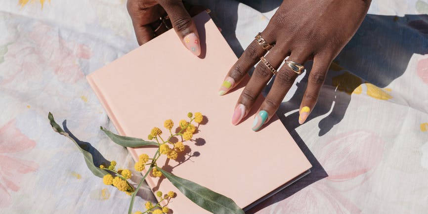 PortraitBlack female hands with rings and colorful nails hold a pastel pink notebook on top of a floral tablecloth, with yellow flowers nearby.of stylish white senior woman wearing black and white clothing and sunglasses, laughing while she holds her pet dogs while seated on a bench at home.